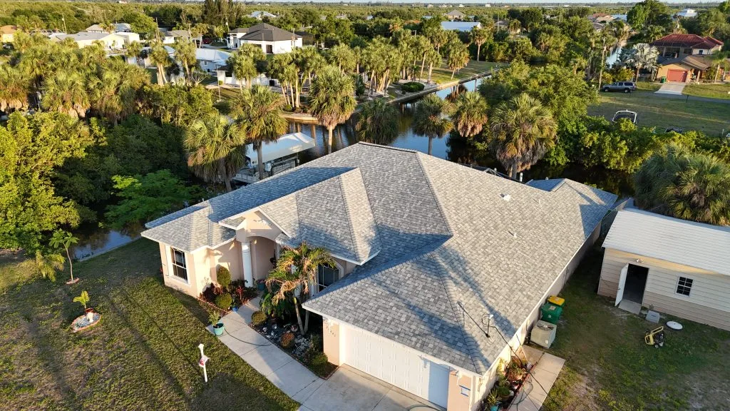 Aerial view of a single-story Florida home with a gray shingle roof, two-car garage, and a canal behind it surrounded by palm trees