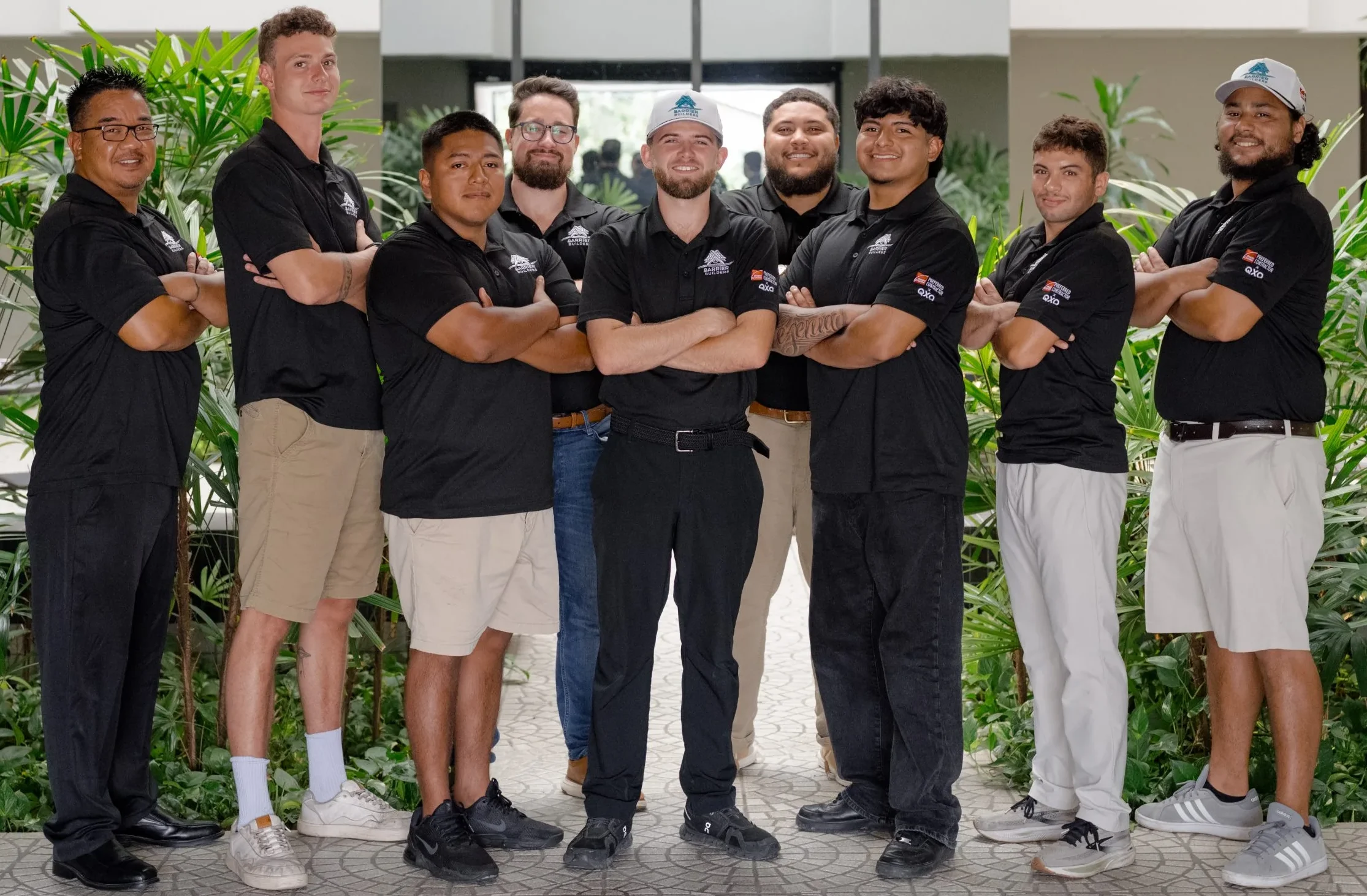 Group of nine Barrier Builders team members in matching black polo shirts posing with arms crossed in a building atrium surrounded by tropical plants